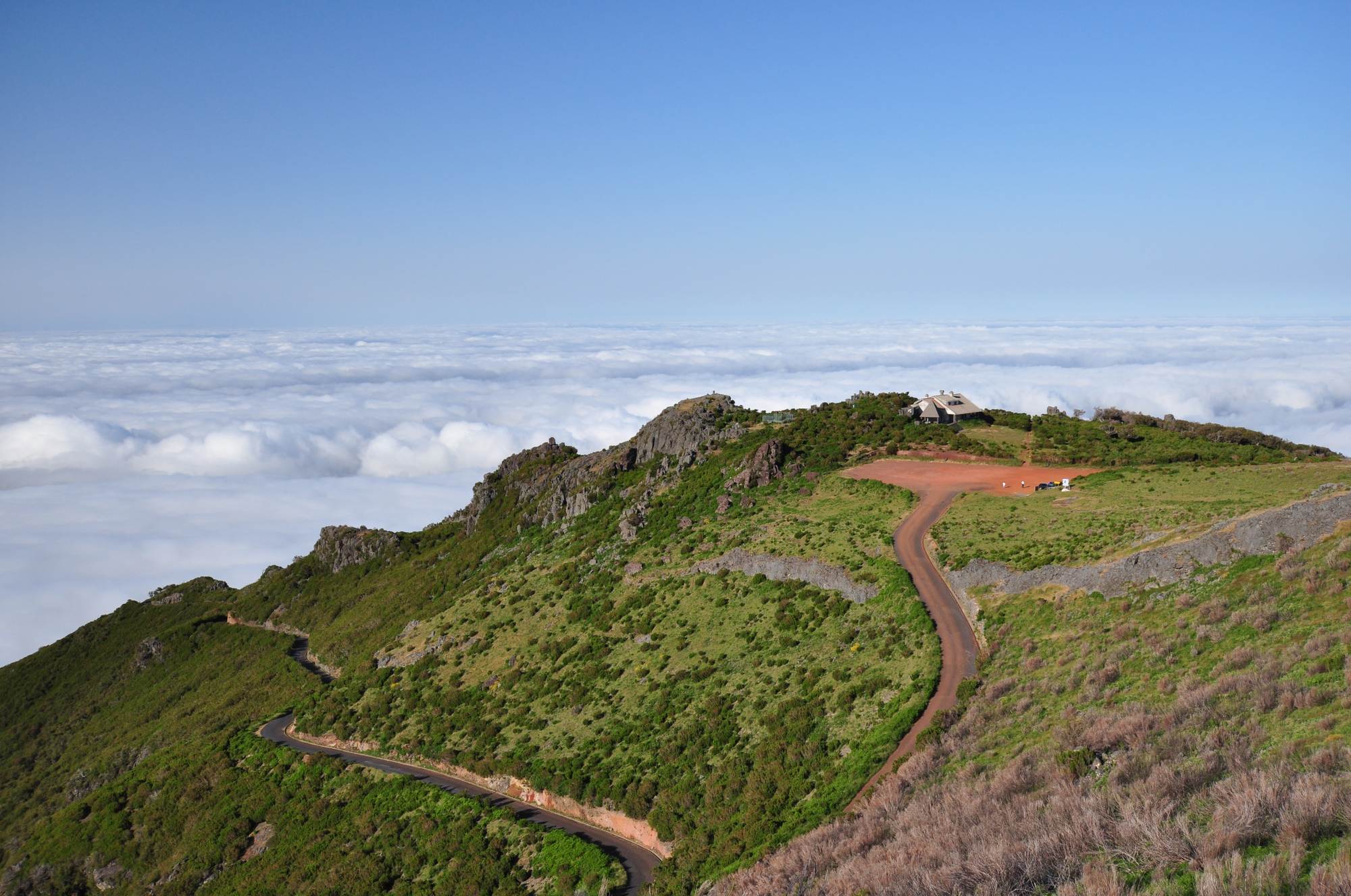 The road climbing to the Achada do Teixeira car park and its stone house, seen from the path to Pico Ruivo, with a layer of cloud below the ridge